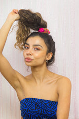 Grainy film image of a model wearing a high ponytail in scrunchie from Uganda. She is sat infront of a pink sparkly background and is wearing a blue strapless tank top